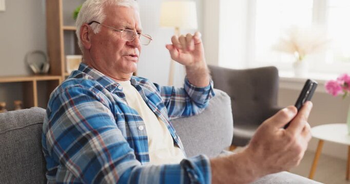 Senior man reading smartphone while wearing glasses. Relaxing at home on a sofa, he focuses on messages, calls and chat technology for conversation. Digital communication in retirement life.