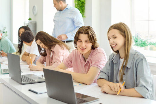 Students teamwork in classroom using laptops for study. Happy teens work on a project, sharing notes and ideas while a teacher guides the group. Emphasizes teamwork and active learning.