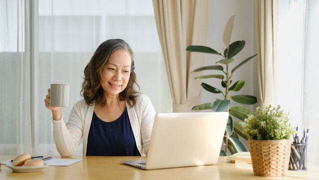 Mature woman relaxing with a cup of coffee while sitting at a home office desk with a laptop in a bright, cozy workspace.