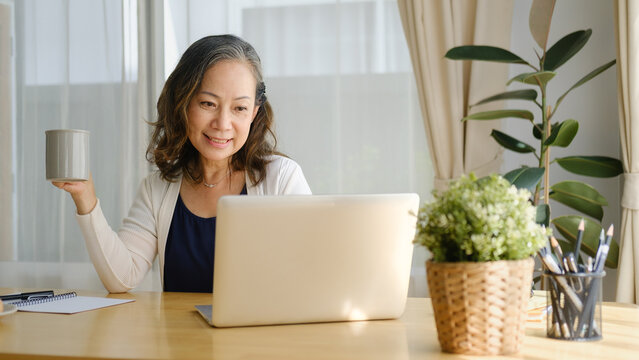 Mature woman relaxing with a cup of coffee while sitting at a home office desk with a laptop in a bright, cozy workspace.