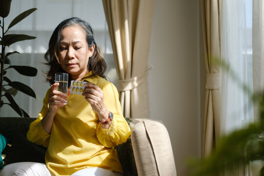 Mature woman holding a glass of water and medicine while sitting at home in natural sunlight. Health care concept.
