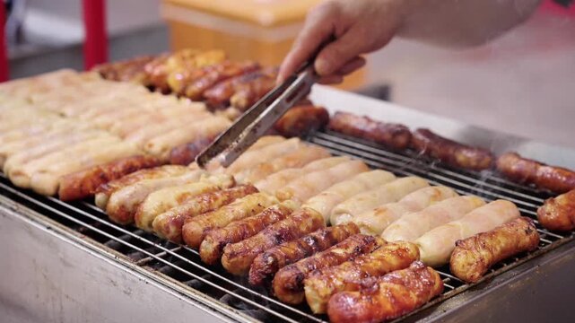 Various sausages are being grilled on a metal grill, with a hand using tongs to handle the food during cooking.