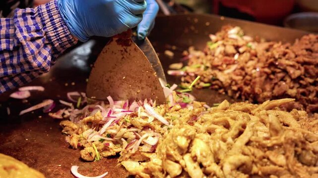 Seasoned sliced meat and red onions being cooked on a flat metal griddle with cooking utensils nearby.