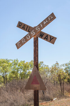 A rusted, vintage metal "Railway Crossing" crossbuck sign stands against a clear blue sky in a dry, rural Australian outback landscape.