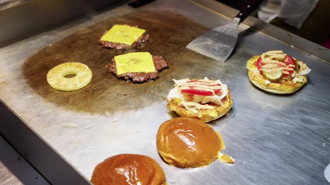 Burger components including beef patties with cheese, pineapple slice, and prepared buns are arranged on a griddle for cooking preparation.
