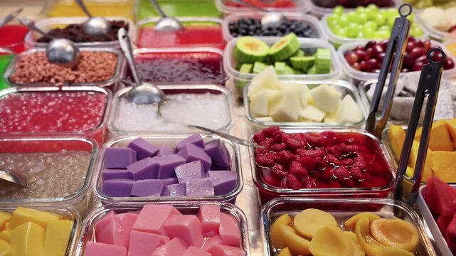 Various colorful dessert toppings, fresh fruits and puddings are displayed in plastic containers for self-service or preparation.