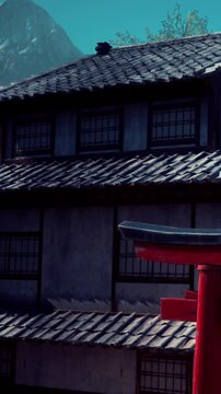 A traditional Japanese building stands next to a red torii gate. The scene shows a well maintained roof and architectural details in a rural setting during the day.