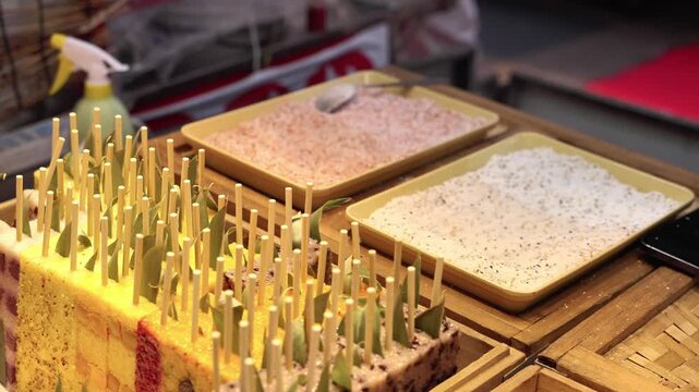 A gloved vendor's hand holds a yellow snack on a stick, preparing it with powder from two trays at a food stall.