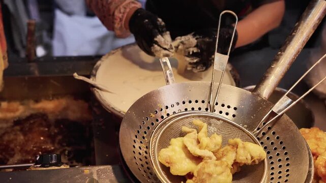 A cook prepares battered food for frying, with fried items in a strainer in a commercial kitchen stall.
