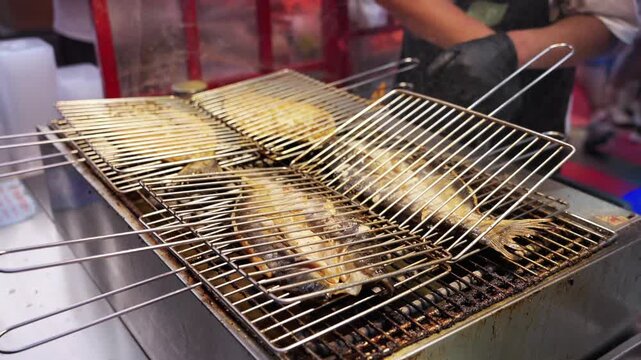 A chef prepares grilled fish using metal grilling racks over a cooking station, wearing black gloves.