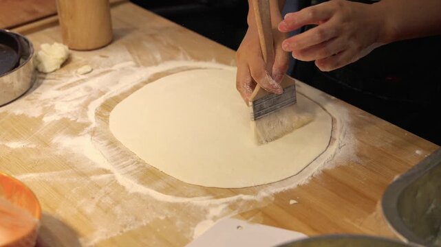 An adult light-skinned person uses a wooden rolling pin to roll flat dough on a floured wooden kitchen counter for baking.