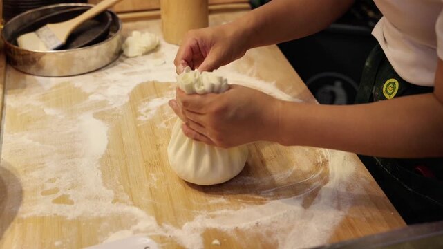 An adult female person folds dumplings with yellow filling on a floured wooden kitchen counter.