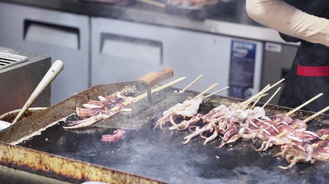 A vendor prepares grilled squid skewers on a hot metal grill, with smoke rising from the cooking food.