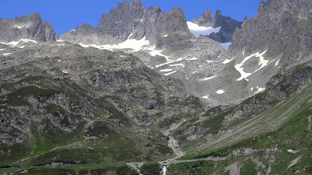 Scenic alpine valley with winding pass road and cascading waterfall amid green slopes and rocky peaks in summer.