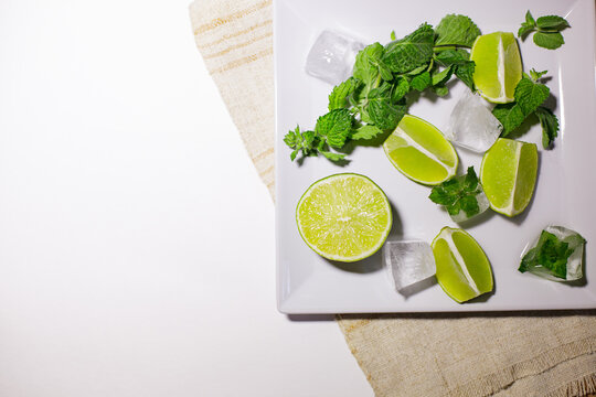 Fresh lime slices, mint leaves and ice cubes on a white plate with large copy space.