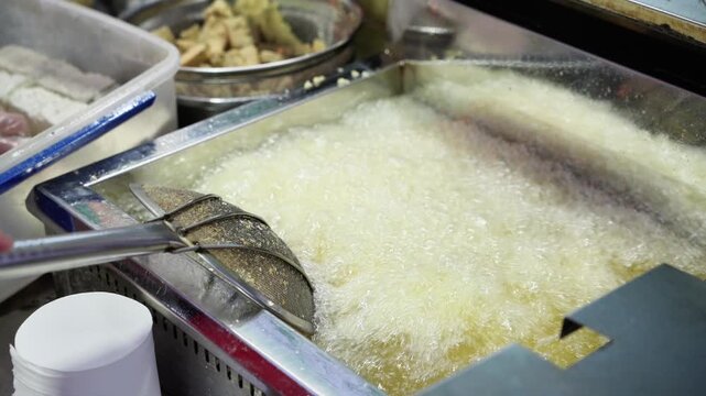 A gloved hand holds raw tofu cubes above a deep fryer of hot oil, with a sieve holding another tofu piece nearby.