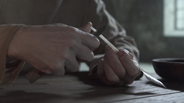 Close up of medieval prisoner hands making a scoop cut on a goose quill shaft with a penknife on a rustic wooden table inside a dark stone dungeon