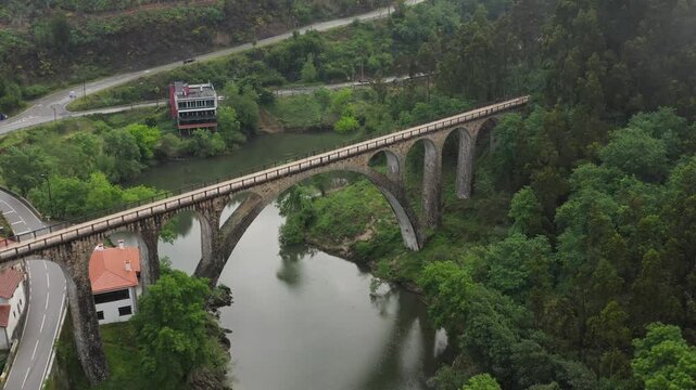 Aerial view of the historic stone arch Poco de Santiago Bridge spanning the river surrounded by lush green forests and winding roads in Sever do Vouga, Aveiro District, Portugal.