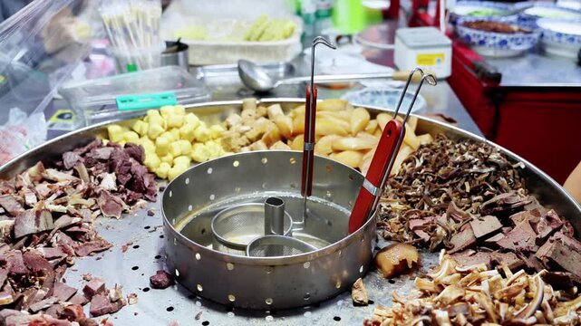 A vendor wearing gloves prepares assorted food ingredients on a metal tray at a food stall, showing various meat and snack items.