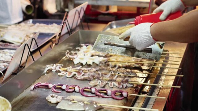 A gloved hand handles skewered squid being grilled on a metal griddle at a street food stall. Fresh seafood skewers are arranged for cooking.