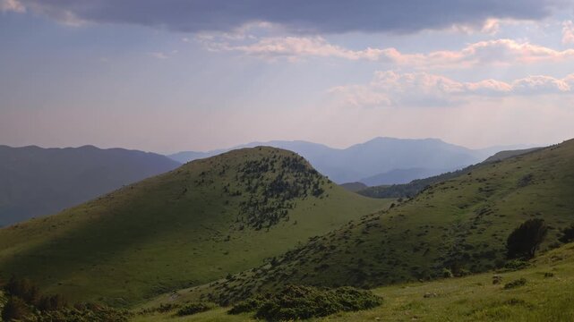 Time-lapse capturing soft evening sunrays filtering through overcast clouds, casting moving shadows and light accents across a beautiful green hill and layered distant mountain ridges during sunset.