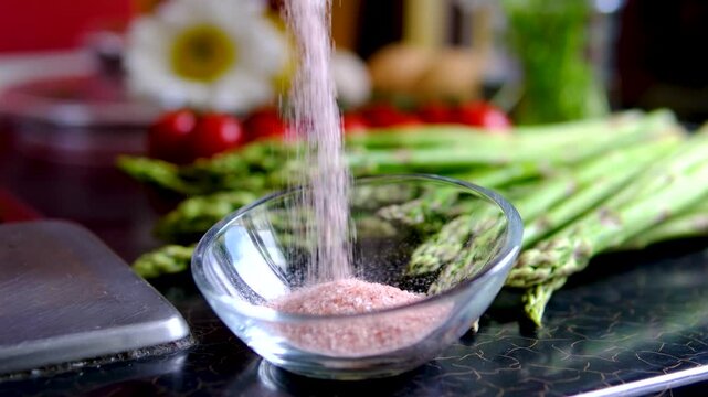 Pouring Himalayan pink salt into glass bowl with fresh asparagus cherry tomatoes and chamomile flowers creating healthy breakfast ingredients for home cooking
