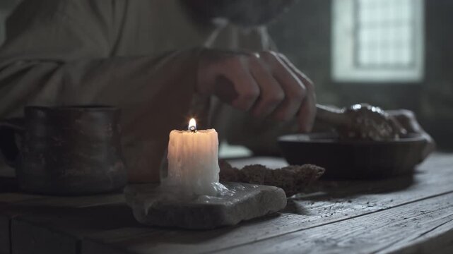 Medieval prisoner eating porridge with a wooden spoon from a bowl near a burning candle and a chunk of rye bread on a rustic table inside a gloomy dungeon, camera moving past