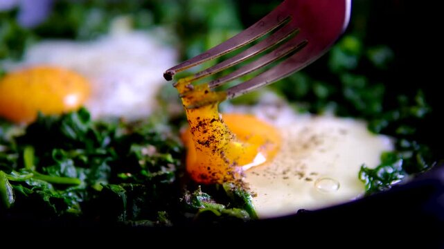 Two fried eggs with runny yolks on spinach in black frying pan seasoned with black pepper as fork breaks the soft yolk for healthy homemade breakfast