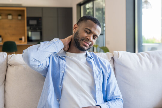 A young man sits on a light sofa at home rubbing his neck in visible discomfort and fatigue, wearing a casual shirt in a bright living room with natural light and modern interior details.