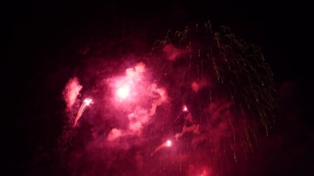 Multiple vibrant fireworks explode in bursts of red and golden light against the black evening sky. Smoke drifts as the festive pyrotechnic show continues