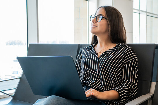 Young woman wearing glasses sitting in an airport lounge, working on her laptop and smiling while looking up, representing travel, connectivity, and future plans