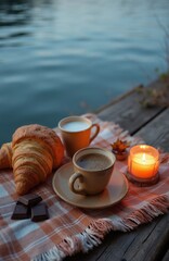 Fototapeta premium Morning coffee and croissant picnic on wooden pier by lake water. Lit candle, chocolate squares add cozy autumn vibe. Relaxing outdoor breakfast by calm blue water.