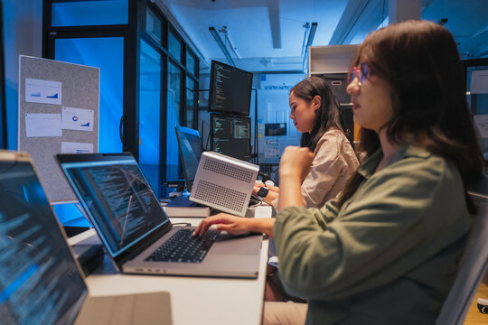A young woman with long auburn hair is seen from behind, typing at a laptop surrounded by multiple monitors displaying lines of code, representing focused solo development work in a tech-driven office