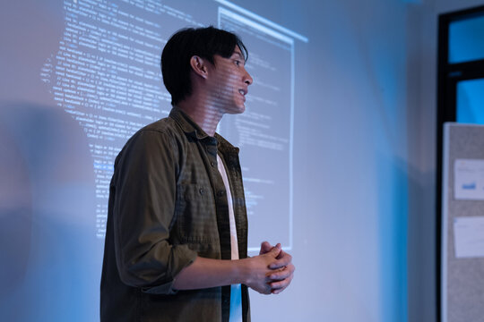 A man gestures toward code displayed on a tablet screen while a seated colleague types on a laptop, surrounded by multiple monitors showing programming code in a blue-lit office.