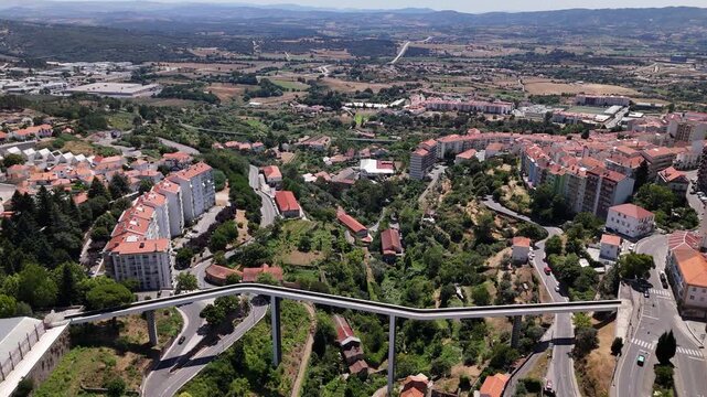 Aerial view of the Carpinteira Pedestrian Bridge spanning the valley in Covilha, with urban buildings, green hills, and winding roads in Covilha, Castelo Branco District, Portugal.