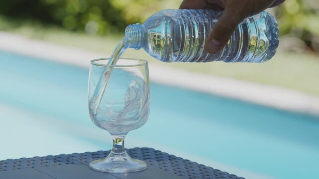 Close-up of fresh clear water being poured from a bottle into an empty glass goblet on a table by a bright blue swimming pool. 