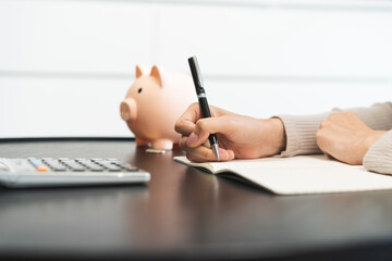 Fototapeta premium Saving money wealth concept, asian young business woman hand write management budget for saving cost, cash finance planning to spend enough money on her income for save money, payment tax, investing.
