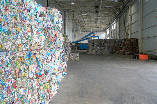 Compressed bales of diverse plastic and paper waste are stacked high inside a vast industrial recycling warehouse, awaiting further material processing