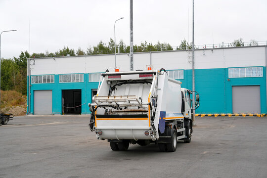 A white garbage truck is parked with its rear compactor open in an outdoor industrial area. A teal and white building stands in the background under a cloudy sky