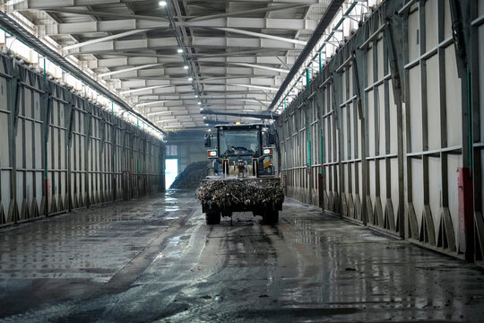 A large vehicle pushes a pile of organic material on a wet concrete floor inside an industrial composting plant. Bright lights illuminate the long processing hall