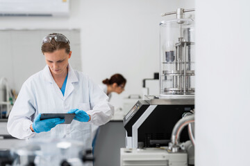 Naklejka premium Technician wears a white lab coat and blue gloves. He holds a tablet, reviewing data. Scientific equipment, including a glass chamber, stands nearby. Another researcher works at a lab station