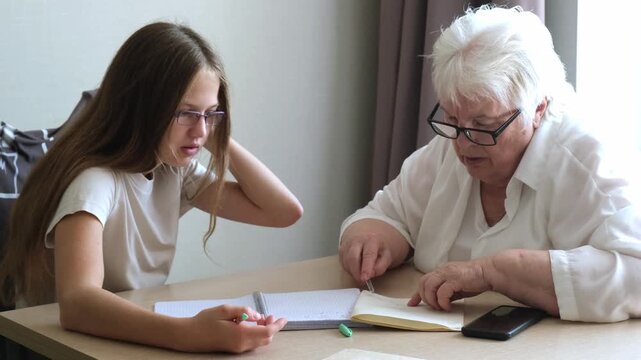 Senior woman and young girl wearing glasses sitting at table doing homework together. Grandmother explaining schoolgirl home tasks