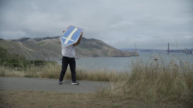 Man Spinning And Tossing Blue Cross Board Overhead And Posing on Coastal Path Bay View
