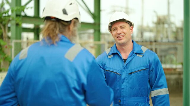 Two professional caucasian male industrial engineers in blue uniforms and hard hat talking, smiling, giving thumbs up at a factory refinery facility. Happy workers discussing project success outdoors