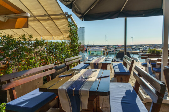 Elegant waterfront restaurant terrace with neatly arranged tables beside a marina filled with boats. Mediterranean hospitality, seaside dining
