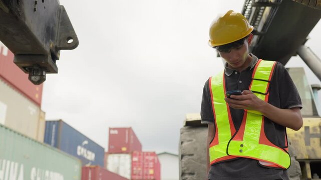 A port worker uses his smartphone for logistics management and to monitor shipping data at a busy container terminal, showcasing modern supply chain technology.