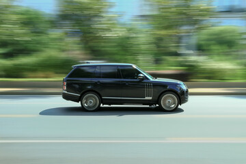 Fototapeta premium Black SUV car driving fast on suburban asphalt roadside with green trees