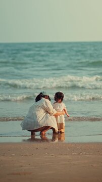 Back view of Asian mother crouching and hugging her little daughter on sandy beach, happy family looking at sea waves during summer vacation for travel tourism, parenting, and childhood lifestyle.