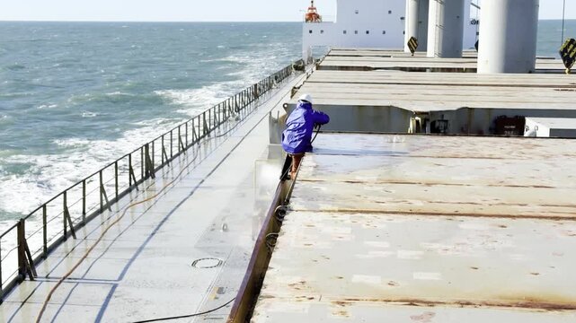seafarer job at sea. vessel crew member in raincoat use high pressure rust chipping machine for ship maintenance.