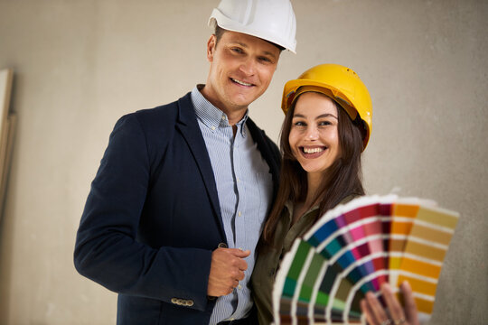 Happy young couple choosing color at construction site and looking at camera.